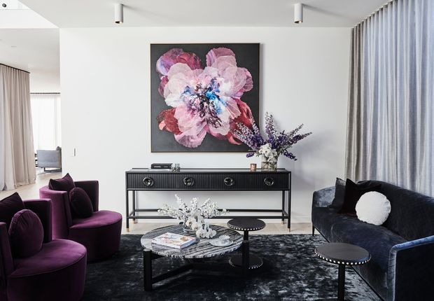 Refined contemporary living room with a dramatic deep-purple and black palette. The space features a dark velvet sofa and sculptural purple armchairs arranged around a round marble-topped coffee table, a black console with decorative floral arrangements, full-height curtains, recessed ceiling lighting, and a large abstract floral artwork. The room is bright but visually moody, with strong contrast and luxe styling.