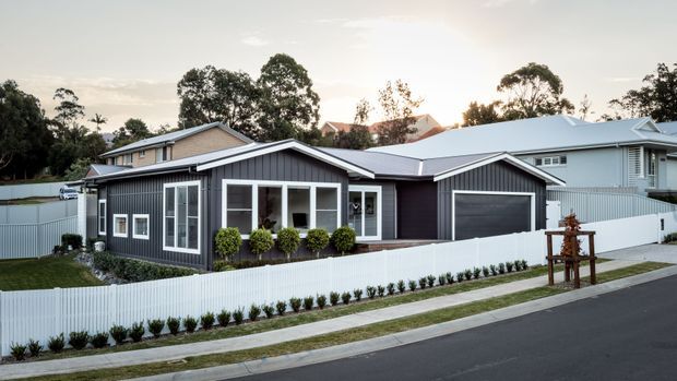 Street-facing view of a contemporary single-storey suburban home with dark vertical cladding, a light metal roof, attached double garage, white picket-style fencing, landscaped planting, and an adjoining covered outdoor area. The house has a low, horizontal form and a clean modern facade.