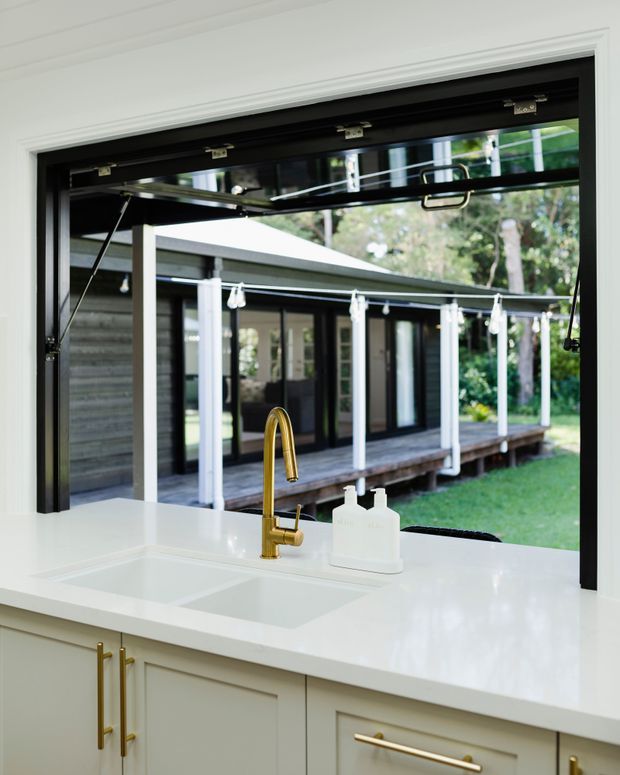 Bright kitchen sink zone photographed from indoors, featuring a white stone-look benchtop, integrated white sink, pale shaker-style cabinetry with long brass handles, and a brass gooseneck tap. A large black-framed awning-style servery window opens toward a covered timber deck and landscaped garden outside.