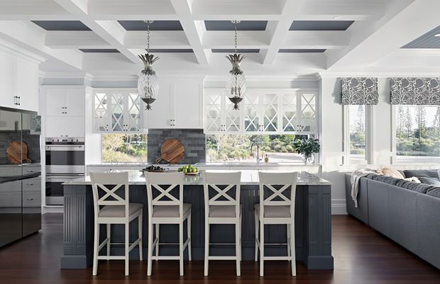 Bright, refined open-plan kitchen with a large dark island and four white X-back bar stools, white cabinetry with glass-front doors, a grey tiled splashback, integrated appliances, and a large window overlooking greenery. The room features a striking coffered ceiling with dark inset panels, decorative pendant lights, dark timber flooring, and an adjacent living area with a sectional sofa.