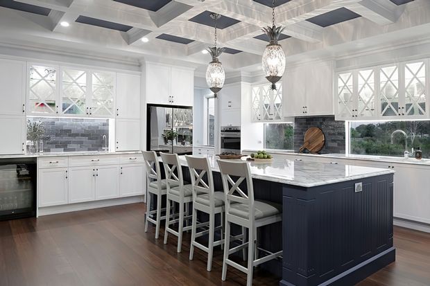 Bright, spacious kitchen with white shaker-style cabinetry, decorative cross-mullioned glass-front doors, a large dark navy island with a pale stone benchtop, grey tiled splashback, timber flooring, and four white bar stools. The coffered ceiling and ornate pendant lights add a refined traditional character.
