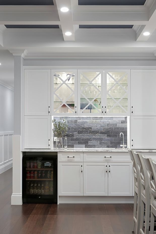 Bright, refined kitchen butler’s-pantry/bar area with white shaker-style cabinetry, glass-front upper cupboards with decorative X mullions, a dark grey stacked-stone tile splashback, integrated sink, beverage refrigerator, and dark timber flooring. The room features substantial traditional moulding and a coffered ceiling with recessed dark panels and downlights. White cross-back stools are visible at the right edge.