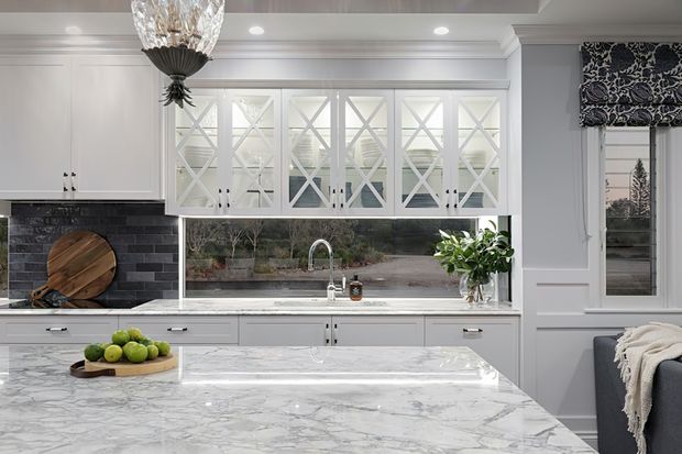 A bright, refined traditional kitchen with white shaker-style cabinetry, glass-front upper cabinets with decorative cross muntins, a dark brick backsplash, marble-look benchtops, and a large central island. A window with a patterned Roman blind brings in natural light, while a faceted pendant light and subtle greenery add decorative detail.