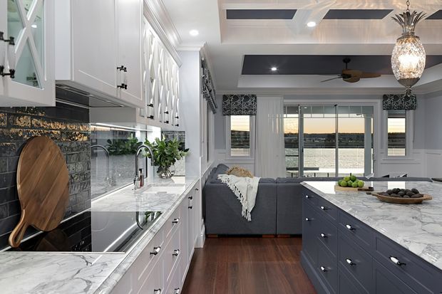 A bright, refined open-plan kitchen flowing into a living area. The kitchen features white shaker-style cabinetry with decorative glass-front details, dark tiled splashback, marble-look benchtops, a large dark island, stainless fixtures, and timber flooring. A grey sectional sofa and waterfront-facing glazing are visible beyond, with patterned window treatments, a decorative pendant light, and a coffered ceiling adding character.