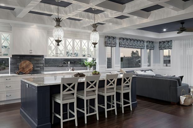 Bright open-plan kitchen and living area with a large dark island, white cabinetry, marble-look benchtop, and four cross-back bar stools. The adjoining living room has a grey sectional sofa and large windows with patterned Roman blinds. Decorative coffered ceiling panels, pendant lights, dark timber flooring, and a ceiling fan add architectural and functional detail.