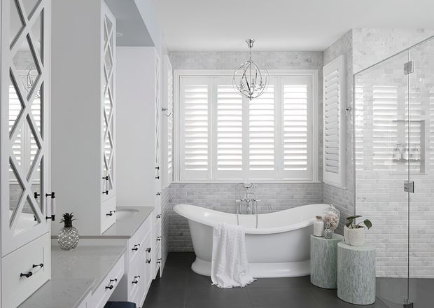 Bright, refined bathroom with a freestanding white pedestal bathtub beneath a spherical chandelier. The room features white shaker-style cabinetry with mirrored panel doors, pale grey engineered-stone benchtops, white plantation shutters, grey-white subway wall tiling, dark flooring, and a frameless glass shower enclosure. Decorative stools and plants add subtle texture.