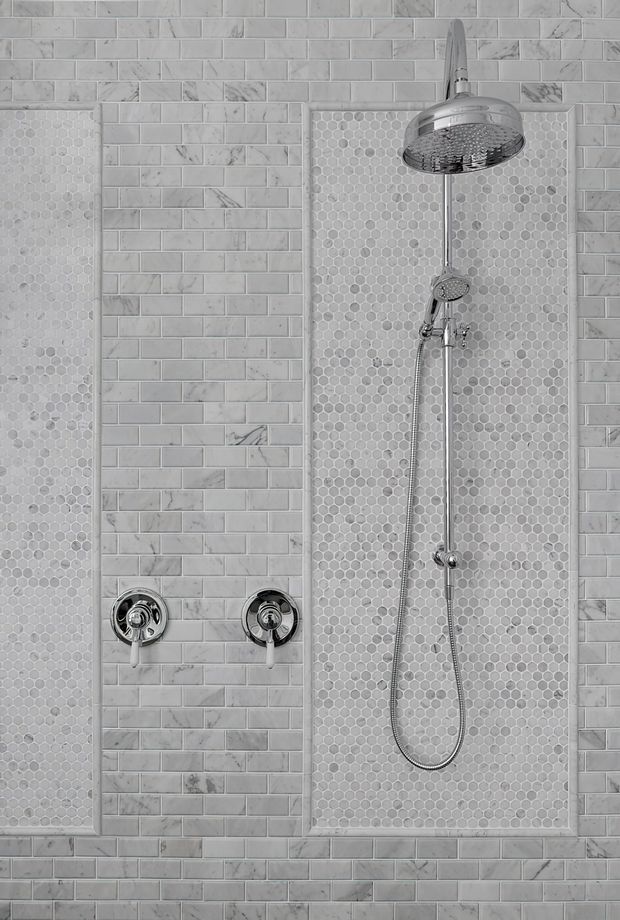 Close-up of a tiled shower wall featuring a large chrome rain showerhead, handheld shower, exposed controls, and a recessed vertical niche. The walls combine white-grey marble-look subway tiles with small hexagonal mosaic tile panels, creating a clean, refined bathroom fit-out.