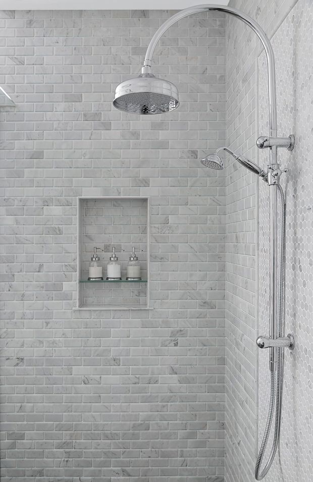 A close-up view of a compact shower enclosure with light grey-white brick-pattern wall tiles, a contrasting small hexagonal tile wall, recessed shelving with soap dispensers, and polished chrome rain shower and handheld fittings.