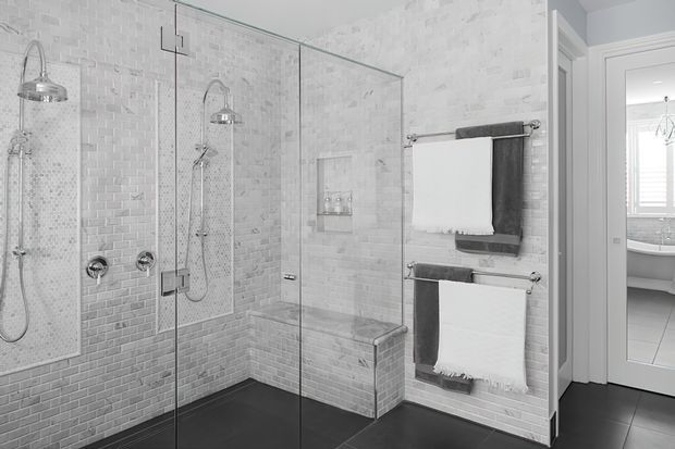 A bright, contemporary bathroom with a large walk-in tiled shower enclosure featuring dual shower heads, handheld fixtures, recessed niches, a built-in tiled bench, and frameless glass panels. White brick-pattern wall tiles contrast with dark floor tiles. Heated towel rails hold white and charcoal towels, and a mirrored door reveals a freestanding bathtub in the adjoining bathroom area.