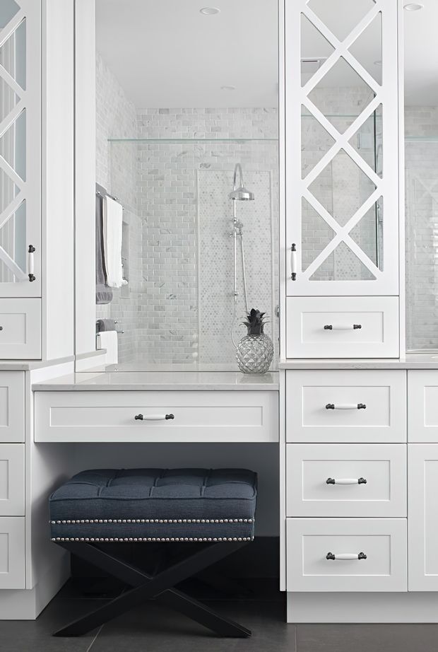 Bright, elegant bathroom vanity area with extensive white built-in cabinetry, mirrored doors with decorative white X-pattern muntins, a pale tiled shower enclosure, glass shelving, and a dark upholstered stool beneath the vanity. The space has a refined monochromatic palette with contrasting dark flooring and hardware.