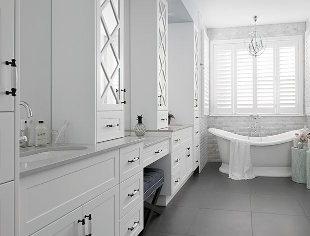 Bright, refined bathroom with extensive white built-in cabinetry, a long vanity and dressing counter, decorative mirrored cabinet doors, grey tiled flooring, and a freestanding white bathtub beneath a large shuttered window. The room has a soft grey-and-white palette with traditional detailing and a delicate pendant chandelier.