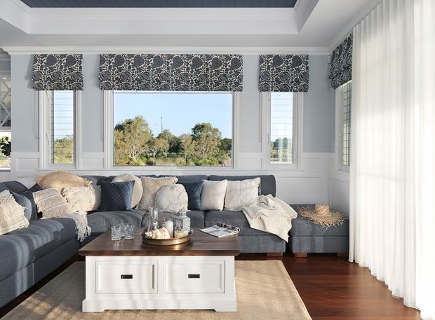 A bright, refined living room with a large charcoal sectional sofa, layered neutral cushions and throws, a dark timber coffee table, and a white storage ottoman. The room features pale grey wall panelling, large windows with patterned Roman blinds, sheer floor-to-ceiling curtains, warm timber flooring, and a recessed coffered ceiling with dark painted boards. Trees are visible outside through the windows.