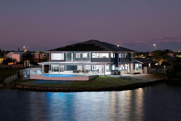 Twilight exterior view of a large contemporary multi-level waterfront residence, with dark pitched roof forms, extensive glazing, balconies and terraces, landscaped lawn, and a brightly illuminated swimming pool facing the canal or lagoon.
