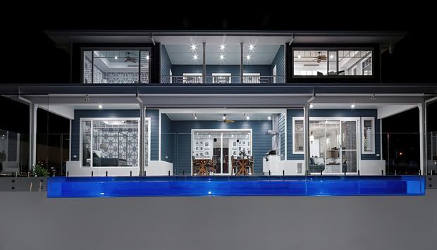 Night-time exterior view of a large contemporary multi-level residence with dark horizontal cladding, extensive white-framed glazing, balconies, and a long illuminated blue swimming pool in front. Interior living and dining spaces are visible through the windows.