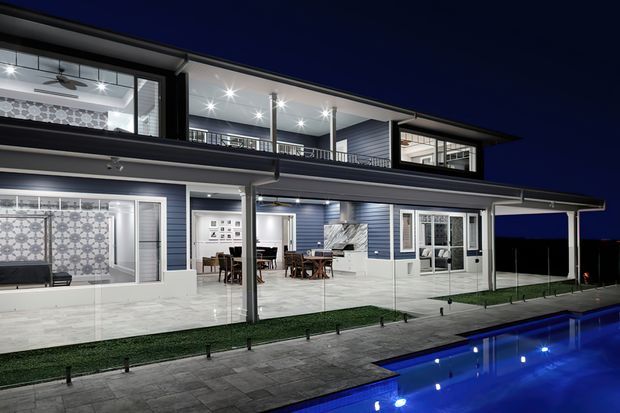 Night-time exterior view of a large contemporary two-storey home with broad roof overhangs, dark and light horizontal cladding, extensive upper-level glazing, covered outdoor entertaining spaces, and a brightly illuminated swimming pool in the foreground. Interior dining and living areas are visible through the glazing, while the main subject is the building facade and poolside setting.