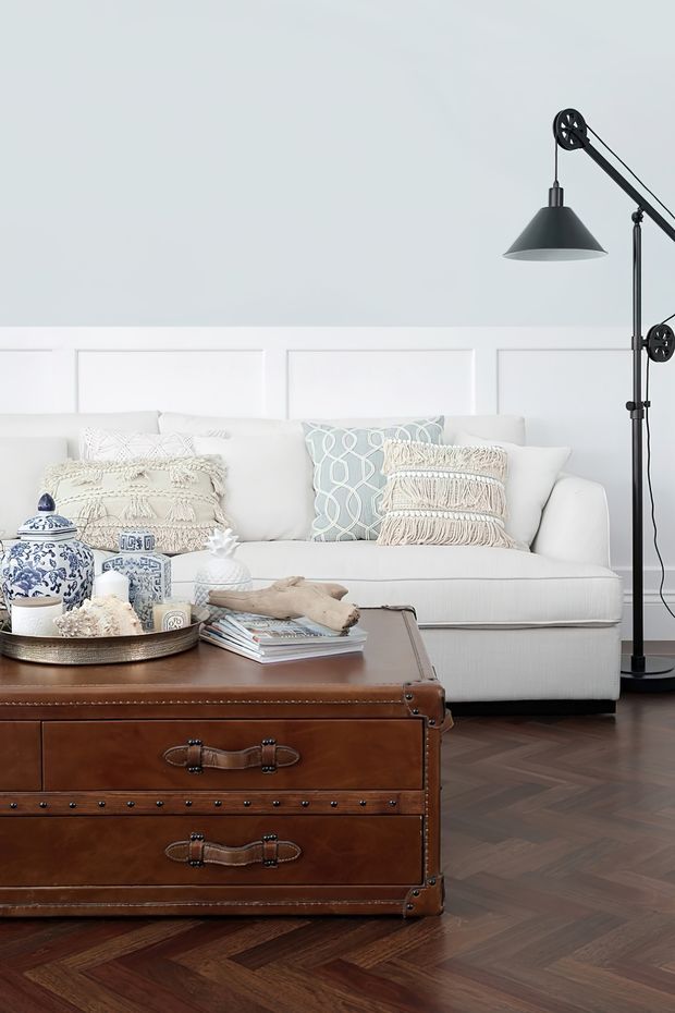 A styled living room featuring a white upholstered sofa with textured cream and pale blue cushions, white decorative wall panelling, a large black articulated floor lamp, and a brown leather trunk-style coffee table. The room has a dark herringbone timber floor and a calm, refined neutral palette with blue decorative accents.