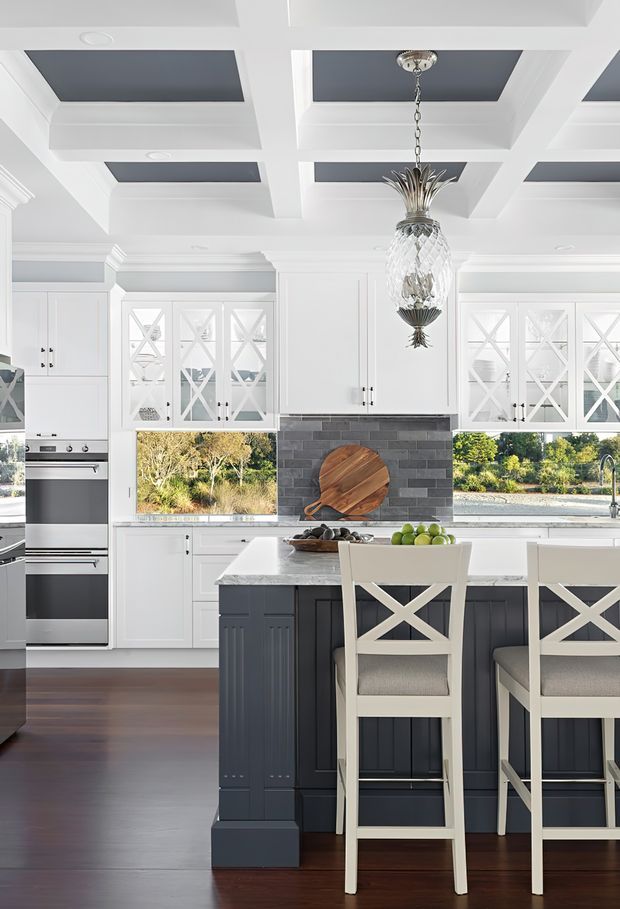 Bright, refined kitchen with white shaker-style cabinetry, glass-front doors with criss-cross muntins, a dark charcoal tiled backsplash, and a large white island with dark paneled cabinetry. The island includes seating for three or more on white cross-back stools. A coffered ceiling with dark inset panels and a decorative crystal pineapple pendant add a traditional, elegant character. Dark timber flooring and views through the windows complete the space.