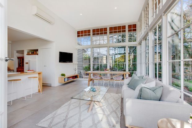 Bright open-plan living room with extensive floor-to-ceiling glazing and white-framed windows, overlooking a garden. The space includes a pale upholstered sofa, glass coffee table, patterned rug, dining table and chairs, and a compact kitchen with island seating. White walls, light grey tiled flooring, and abundant natural light create an airy contemporary interior.