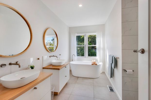 Bright, spacious contemporary bathroom with two timber-framed round mirrors, twin white vessel basins on timber-look vanity tops, a freestanding white bathtub, pale tiled flooring, white walls, and a large window overlooking greenery. Brushed metal tapware and fixtures provide subtle contrast.