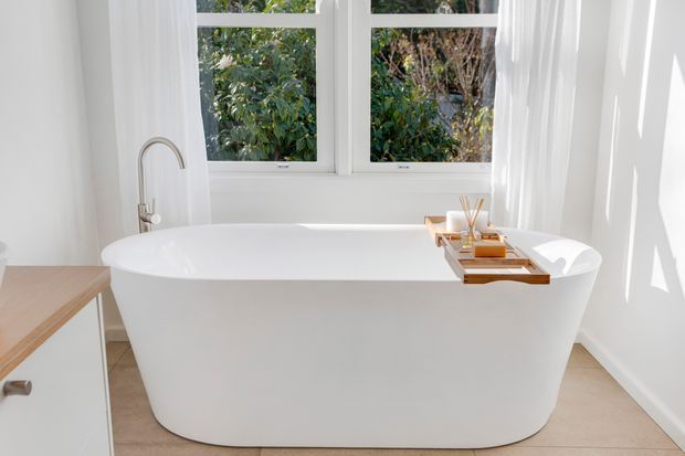 Bright, serene bathroom featuring a freestanding white oval bathtub positioned beneath large white-framed windows, with a wall-mounted or deck-mounted brushed metal bath filler at the left. A timber bath caddy with toiletries spans the tub, while sheer curtains, pale tiled flooring, and white walls create an airy minimalist setting.