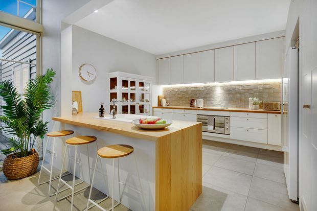 Bright contemporary kitchen with a large light timber island and three white bar stools, white full-height cabinetry, integrated appliances, a textured stone-look backsplash with under-cabinet lighting, pale tiled flooring, and a glazed opening with indoor plants. A small dining or living area is partially visible beyond the kitchen.
