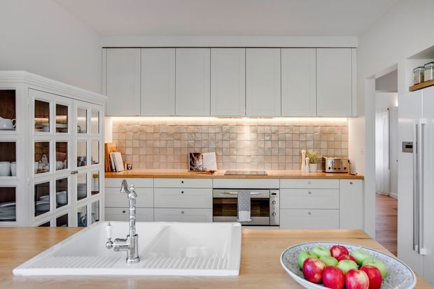Bright, refined kitchen with white floor-to-ceiling cabinetry, glass-front display cupboards, a timber benchtop, large white farmhouse-style sink, stainless steel tapware and appliances, and a warm beige tiled splashback illuminated by under-cabinet lighting. A central island occupies the foreground, with a hallway visible beyond the kitchen.
