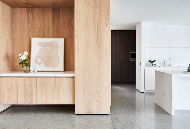 A refined open-plan interior showing a minimalist kitchen and built-in timber joinery. Pale natural timber cabinetry and wall panelling frame a recessed display niche, while a large pale stone island and white kitchen surfaces occupy the right side. The polished grey terrazzo or concrete floor, concealed appliances, and clean-lined detailing create a calm, contemporary space.