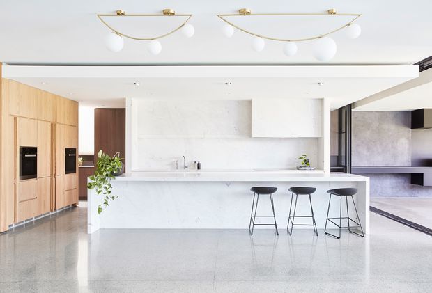 Bright open-plan contemporary kitchen with a long white stone island and three black bar stools. Light timber floor-to-ceiling cabinetry with integrated appliances lines the left wall, while a white marble-look splashback and cabinetry form the main kitchen backdrop. Polished grey concrete or terrazzo-style flooring extends into an adjacent open living area, and sculptural brass-and-white ceiling lights add a refined accent.