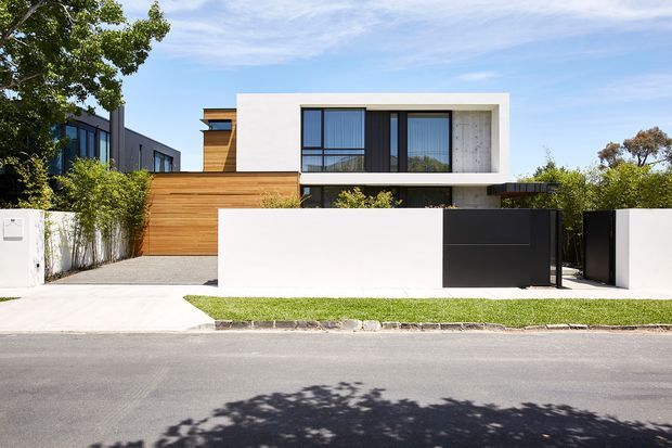 Street-facing view of a contemporary two-storey residence with a crisp white rectangular upper volume, extensive dark-framed glazing, warm horizontal timber cladding, and a dark vehicle gate. The property has a minimalist front boundary wall, concrete driveway and landscaped planting in a suburban setting.