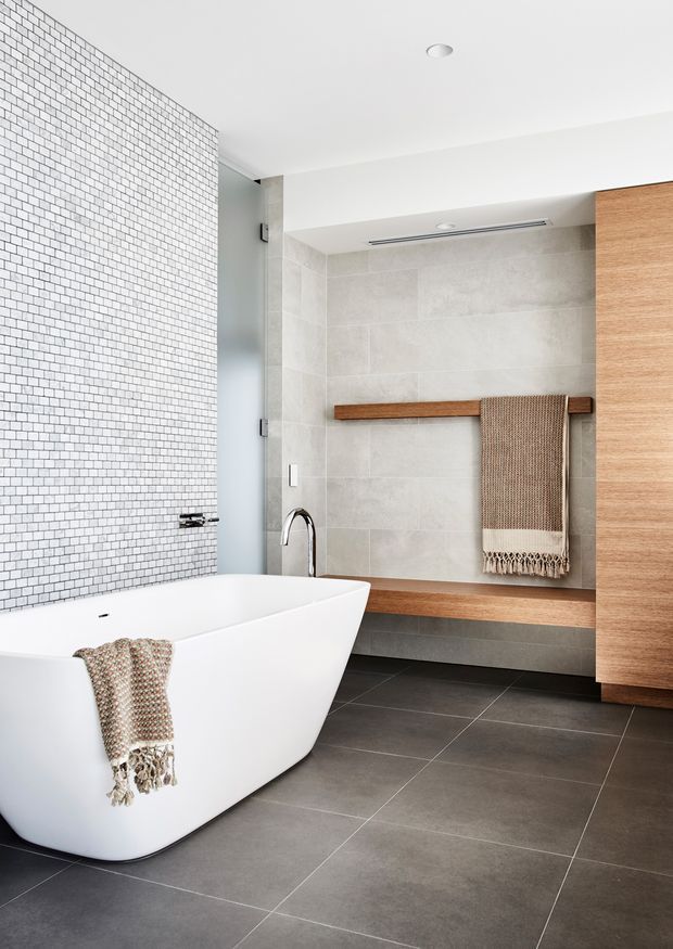 Bright contemporary bathroom featuring a freestanding white bathtub, a walk-in shower with pale large-format wall tiles, a small white mosaic-tile feature wall, dark grey floor tiles, warm timber bench and towel rail, and tall timber cabinetry. Minimal recessed lighting and clean-lined fixtures create a refined, spa-like setting.