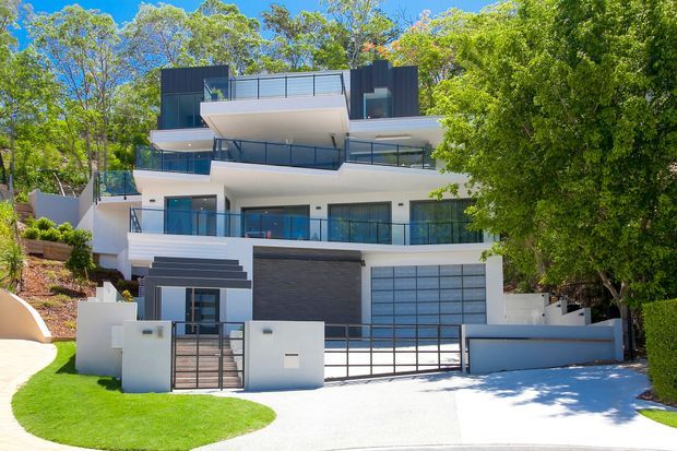 A contemporary multi-storey residence set among mature trees on a sloping suburban site. The facade combines white rendered walls with dark charcoal cladding, extensive glass balustrades and glazing, projecting balconies, and a large integrated garage door. A landscaped lawn, driveway, entry gate, and retaining walls are visible at the front.