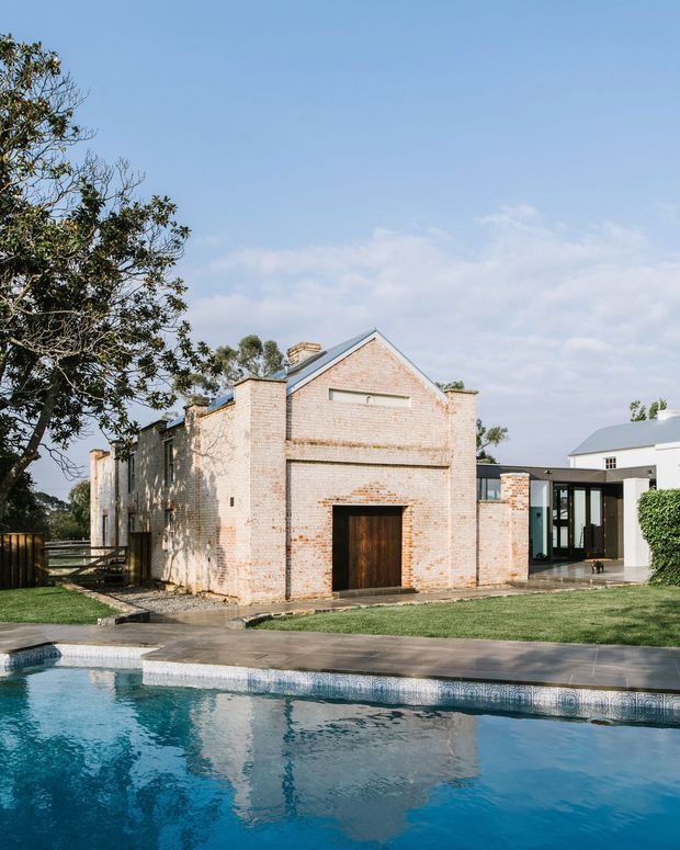 Exterior view of a renovated brick building beside a swimming pool. The main structure has pale exposed brick walls, a simple pitched gable roof with metal roofing, timber garage-style doors, and a modern glazed link connecting it to a crisp white contemporary building. A paved pool edge, lawn, mature trees, and hedges complete the residential setting.