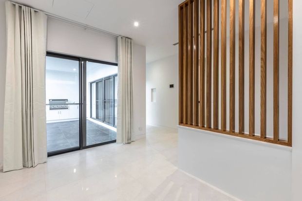 Bright, minimalist interior transition space with glossy pale flooring, white walls and ceiling, full-height curtains, and large black-framed sliding glass doors opening to a tiled outdoor courtyard or terrace. A prominent vertical timber slat screen provides partial separation and warmth on the right.