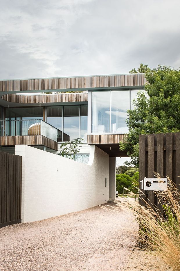 Exterior view of a contemporary multi-level residence from a gravel driveway or entry path. The building features extensive floor-to-ceiling glazing, dark metal window frames, curved and projecting forms, white masonry walls, and prominent vertical timber cladding. A dark timber boundary fence, landscaped planting, and a covered passage beneath the upper level are visible.