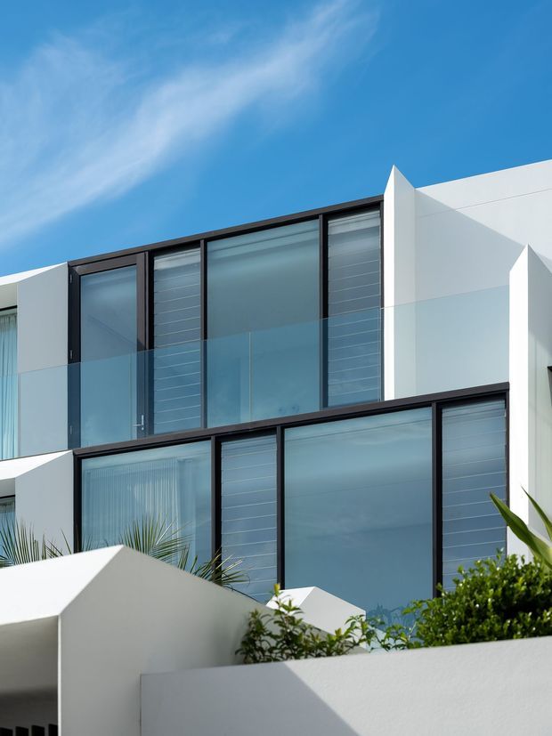 Exterior view of a contemporary multi-level residence with crisp white geometric walls, expansive dark-framed glazing, translucent glass balustrades, and projecting angular roof or parapet forms. Tropical planting is visible in the foreground beneath a clear blue sky.