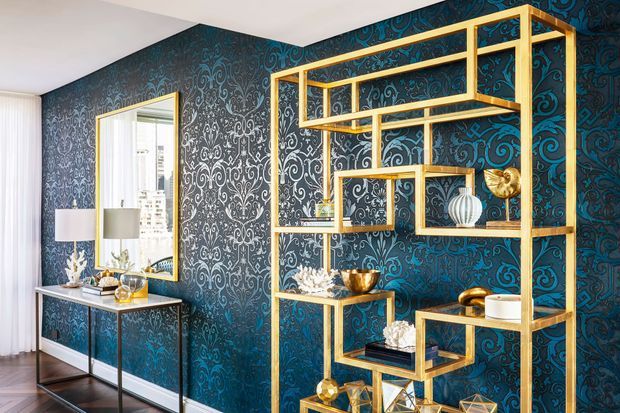 A refined interior vignette featuring a large geometric gold-toned open shelving unit against dramatic dark teal and black damask wallpaper. Decorative objects, a console table, mirror, table lamps, and sheer curtains create a polished, glamorous look, with dark timber flooring below.