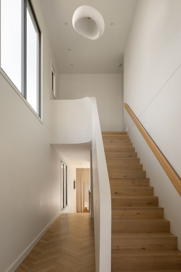 A bright, minimalist interior stairway with warm timber stairs and matching handrail ascending along a white wall. A sculptural curved white balustrade encloses the stair opening, while large vertical glazing brings in natural light. The lower level continues into a hallway with timber flooring and a softly curtained room beyond. A simple sculptural ceiling light and recessed downlights complete the clean, restrained design.