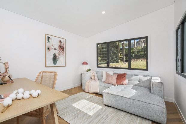 Bright, compact open-plan living and dining area with white walls, a pale timber dining table and woven dining chairs, a light grey sectional sofa, neutral rug, black-framed windows, and understated pastel artwork and cushions. The room has a clean, relaxed contemporary feel with abundant natural light.