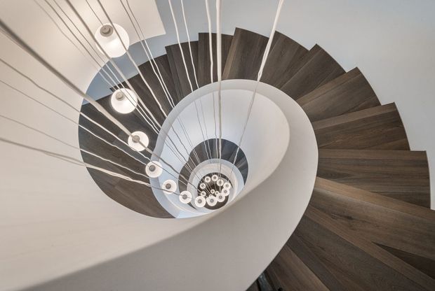 Dramatic upward view through a sculptural spiral staircase, featuring dark timber treads curving around a white central void and a cascading cluster of suspended pendant lights. The composition emphasizes the geometric spiral, contrasting materials, and strong architectural form.