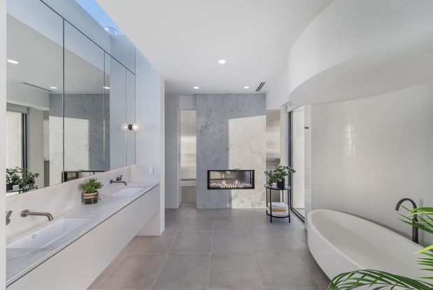 Bright contemporary bathroom with a long double vanity and mirrored wall on the left, a freestanding curved bathtub on the right, large grey floor tiles, pale textured wall finishes, and a central linear fireplace set into a stone-look feature wall. Recessed lighting and curved ceiling detailing create a clean, luxurious feel.