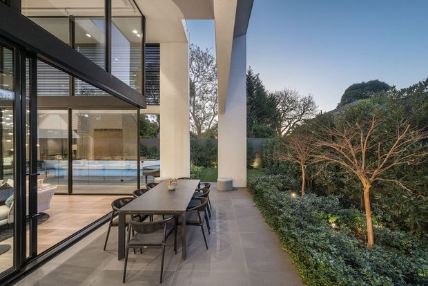 A contemporary covered outdoor dining terrace beside a modern glass-walled residence. A long dark dining table with black chairs sits on large grey pavers, framed by tall white structural columns and expansive black-framed glazing. The glazing reveals a bright interior and reflects parts of the terrace, while a dense landscaped garden borders the space.