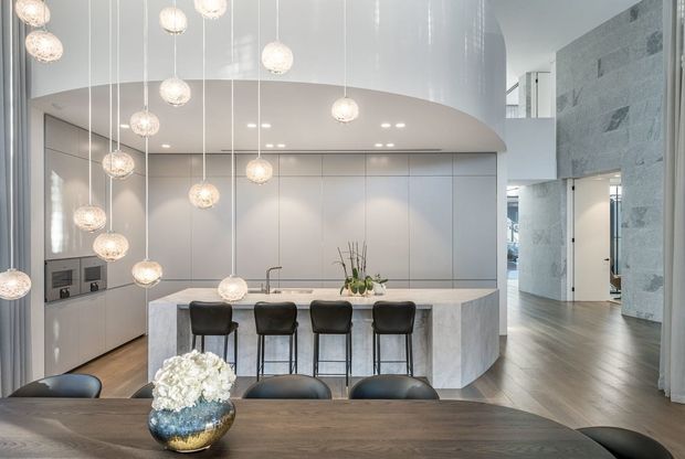 A refined open-plan kitchen and dining area with a large pale stone island, integrated light cabinetry, four dark bar stools, and a timber dining table in the foreground. Numerous spherical pendant lights hang at different heights beneath a curved white ceiling feature. The space opens toward a tall, light grey stone-clad wall and adjoining circulation area, creating a bright, polished contemporary interior.
