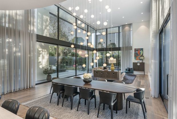A sophisticated double-height dining and living area with extensive floor-to-ceiling glazing, sheer curtains, dark-framed windows, and a dramatic cluster of spherical pendant lights. A long oval timber dining table with dark upholstered chairs sits on a large rug in the foreground, while a sideboard, lounge seating, artwork, and indoor planting occupy the living zone beyond. The space is bright, refined, and contemporary, with views into a landscaped courtyard.