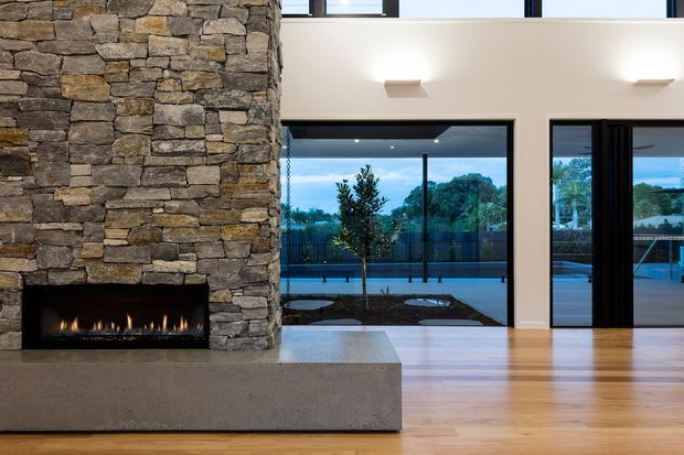 Contemporary double-height living space featuring a substantial floor-to-ceiling rough-stacked stone fireplace with a linear firebox, polished concrete hearth, warm timber flooring, and expansive dark-framed glazing opening to a pool terrace. A mezzanine-level window and simple wall-mounted lighting contribute to the airy, modern composition.