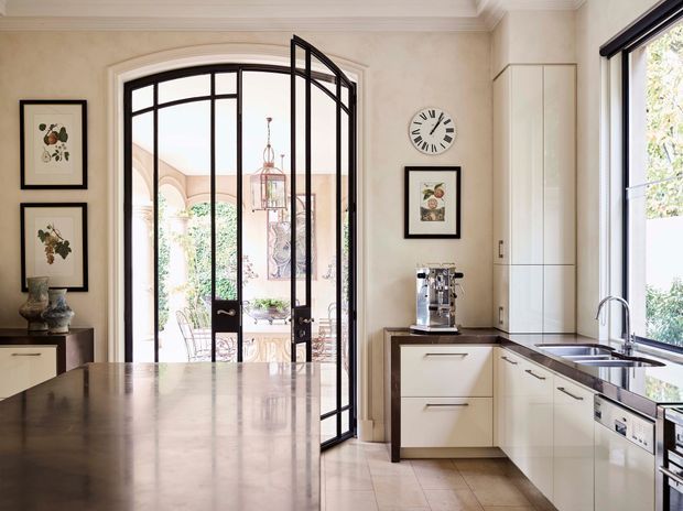A bright, refined kitchen with a large stone island in the foreground, pale built-in cabinetry, dark stone benchtops, and a stainless-steel sink and dishwasher. A dramatic black-framed glazed French door opens to a courtyard or outdoor area, while framed botanical artwork, a wall clock, and a coffee machine add detail. The room has a warm neutral palette and traditional architectural detailing, including an arched opening and decorative cornice.