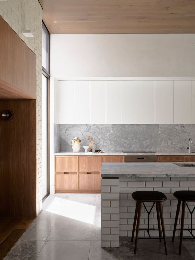 A refined contemporary kitchen with a large island and two dark timber stools. The room features white tiled walls, pale timber-lined ceiling, white upper cabinetry, warm timber lower cabinetry, a grey veined stone splashback and benchtop, and large pale floor tiles. A dark timber built-in wall niche and tall window add depth and natural light.
