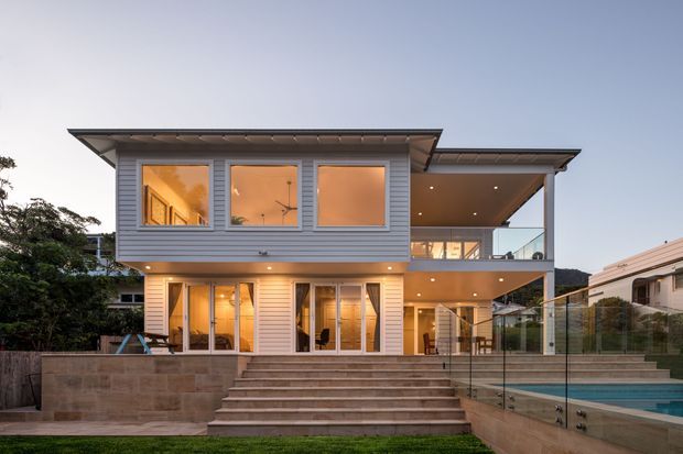 A contemporary two-storey suburban home viewed from the rear, featuring white horizontal weatherboard cladding, broad glazed openings, a sheltered upper-level terrace, and extensive outdoor steps leading to a swimming pool. Warm interior lighting contrasts with the cool dusk exterior.
