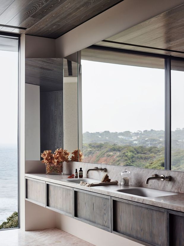 A refined contemporary bathroom vanity overlooking a coastal landscape. The room features a long double-sink vanity with dark stained timber cabinetry, a pale stone or marble benchtop and integrated backsplash, wall-mounted brushed metal tapware, a large frameless window, and dark timber ceiling panels. The space is calm and minimalist with strong natural light and expansive ocean and greenery views.