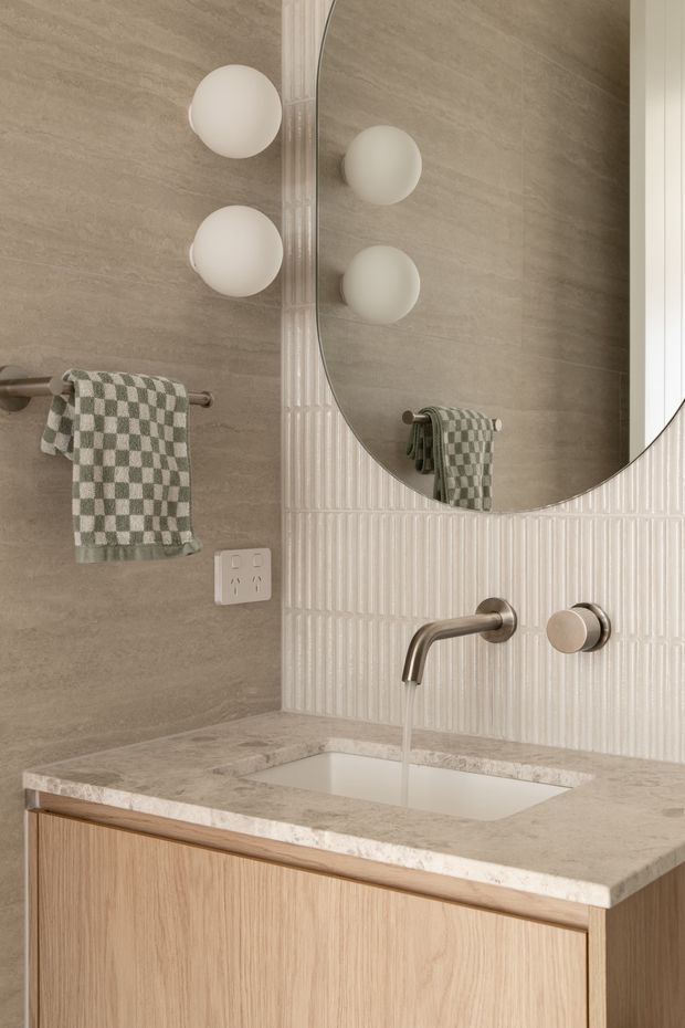 Close-up view of a refined contemporary bathroom vanity. The scene features a light timber floating vanity with an integrated white basin, pale stone benchtop, brushed bronze wall-mounted tapware, warm grey stone-look wall tiles, and a vertically ribbed cream tile splashback. A large rounded mirror reflects globe wall lights and a checked hand towel on a simple metal rail.