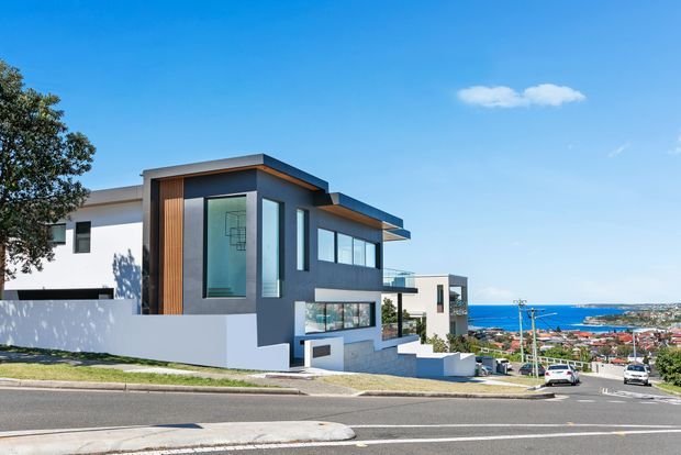 Street-level exterior view of a contemporary multi-level residence on a sloping coastal suburban site. The home features crisp white rendered walls, dark grey geometric volumes, warm vertical timber cladding, extensive glazing, projecting flat roof planes, and balconies. The ocean, bay, and surrounding suburban settlement are visible beyond the property.